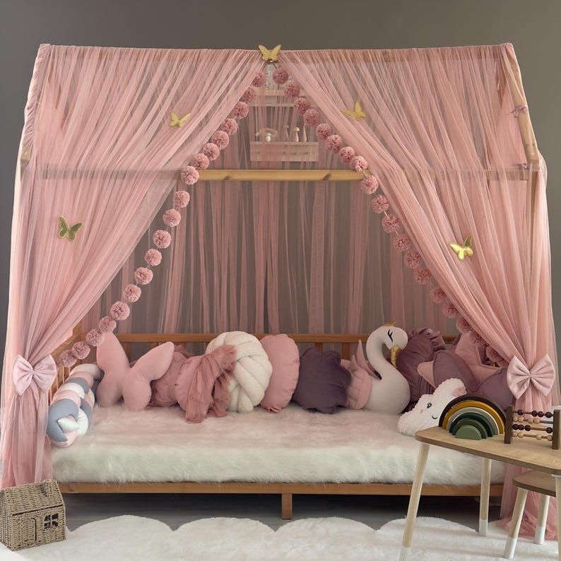 May include: A pink canopy bed with sheer curtains, adorned with butterfly accents and pom-pom garlands. The bed is furnished with various decorative pillows, including butterfly and swan-shaped ones, and sits on a fluffy white rug.