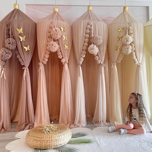 Puede incluir: Cuatro cortinas de dosel beige transparentes con ribete de pompones y detalles de mariposas doradas. Una ni&ntilde;a est&aacute; sentada en una alfombra blanca frente a las cortinas.