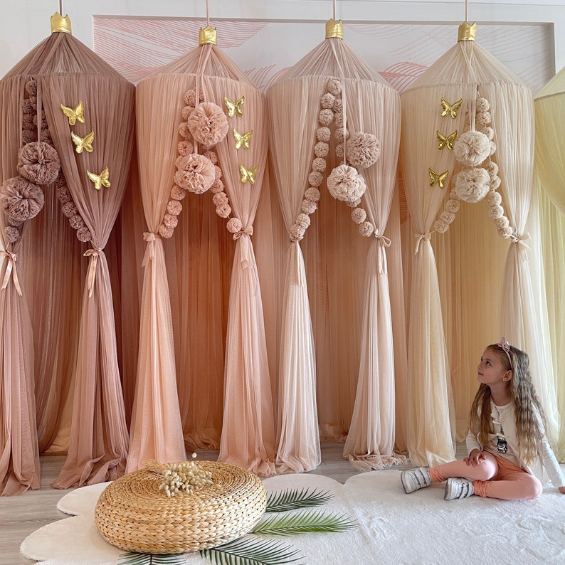 Puede incluir: Cuatro cortinas de dosel beige transparentes con ribete de pompones y detalles de mariposas doradas. Una ni&ntilde;a est&aacute; sentada en una alfombra blanca frente a las cortinas.