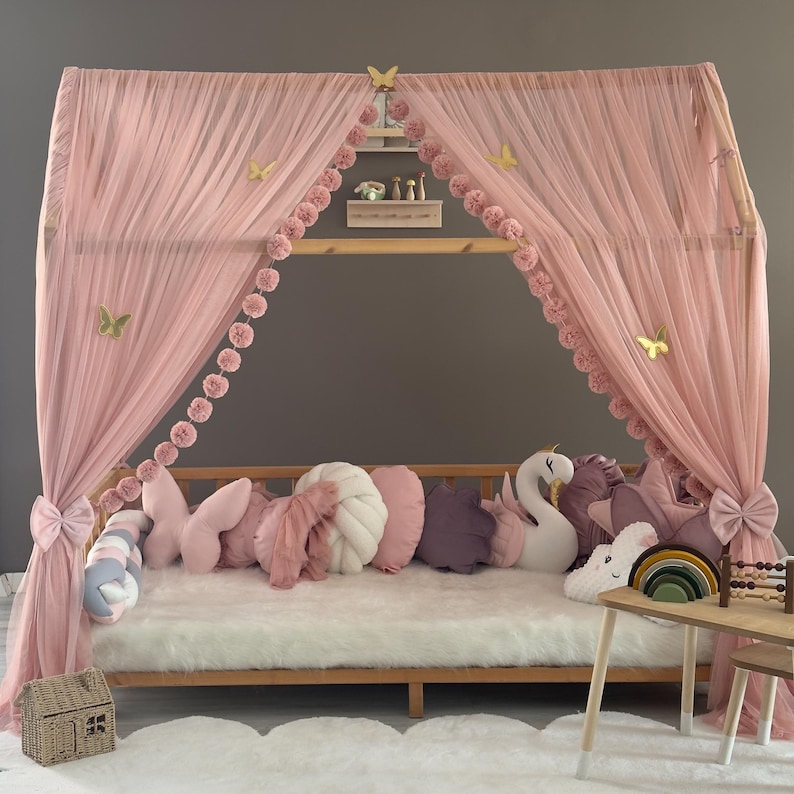 May include: A wooden canopy bed draped with pink tulle and decorated with butterfly accents. The bed is adorned with various decorative pillows in shades of pink, white, and purple. A small wooden table with toys sits nearby.