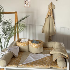 Puede incluir: Un juego de cambiador para bebés con un estampado floral amarillo y blanco. El juego incluye un cambiador, dos cestas de almacenamiento, una toalla y una almohada. El cambiador está rodeado por un parachoques beige. Una ilustración de león enmarcada cuelga de la pared.