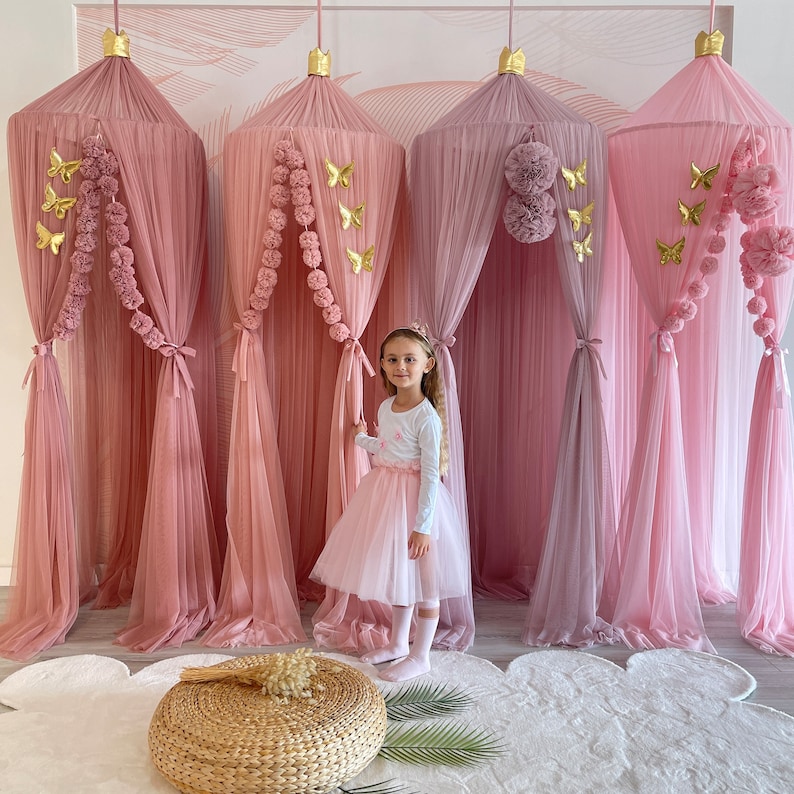 May include: Four pink tulle canopy beds with gold butterfly decorations and pom pom trim. A young girl is standing in front of the canopies wearing a pink tulle skirt and a white long-sleeved shirt.