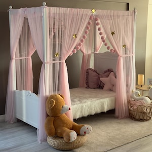 May include: A white canopy bed draped with sheer pink fabric and adorned with gold star accents. The bed is decorated with pink pillows and a star-shaped pillow. A large teddy bear sits beside the bed on a woven stool. A basket with a swan figurine is also present.