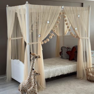Pode incluir: Uma cama com dossel branca com cortinas transparentes creme e decorações em forma de estrela. A cama é decorada com um cobertor branco fofo e almofadas. Duas girafas de pelúcia estão em frente à cama.