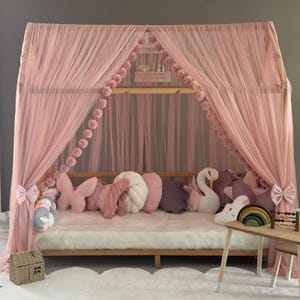 May include: A pink canopy bed with sheer curtains and decorative pom-poms. The bed is adorned with various pillows in shades of pink, white, and purple. A small wooden table with a rainbow and abacus sits nearby, creating a cozy children's bedroom setting.