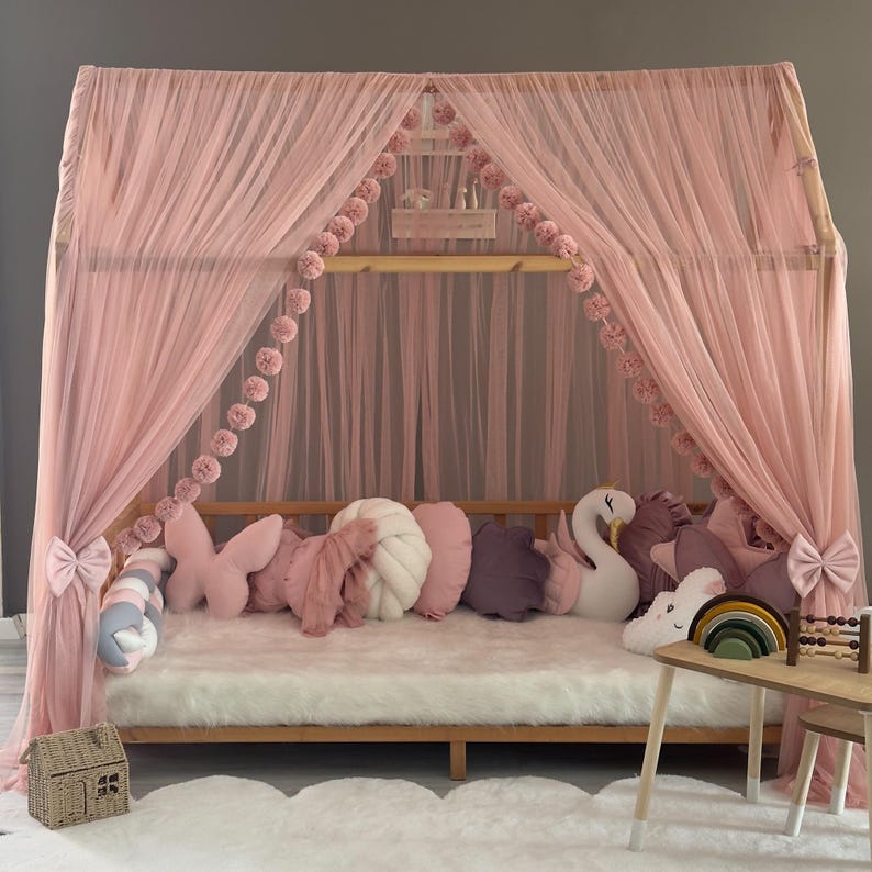 May include: A pink canopy bed with sheer curtains and decorative pom-poms. The bed is adorned with various pillows in shades of pink, white, and purple. A small wooden table with a rainbow and abacus sits nearby, creating a cozy children's bedroom setting.