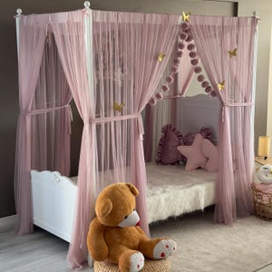 Pode incluir: Uma cama branca com dossel rosa e cortinas transparentes. A cama é decorada com almofadas rosa e uma almofada em forma de estrela. Um grande urso de pelúcia marrom está sentado em um banquinho de vime em frente à cama. Decorações de borboletas douradas estão presas ao dossel.