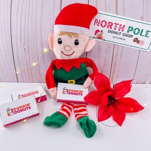 May include: A plush elf doll wearing a red hat and green and red outfit, holding a Dunkin' Donuts gift card. A sign that reads "North Pole Donut Shop" is in the background, along with a red poinsettia and small Dunkin' Donuts boxes.