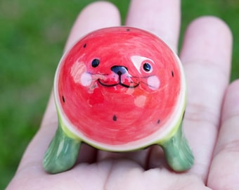 May include: A ceramic figurine of a seal shaped like a watermelon. The seal has a red and green watermelon body with black eyes and a smiling mouth.