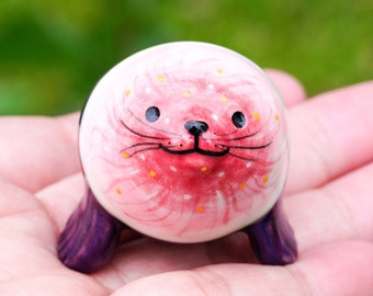 May include: A small ceramic figurine of a seal with a pink and white body and purple flippers. The seal has a painted face with black eyes, a red nose, and a yellow mouth.