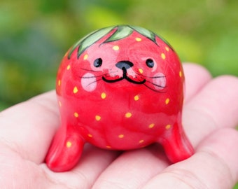 May include: A ceramic figurine of a strawberry with a cute cartoon face. The strawberry is red with yellow seeds and green leaves.