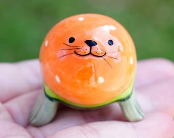 May include: A small ceramic seal figurine with a bright orange body, white spots, and a green and gray base. The seal has a painted face with black eyes, a black nose, and a black mouth.