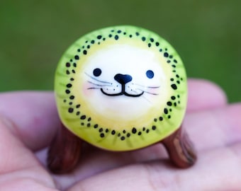 May include: A small ceramic figurine of a seal wearing a kiwi fruit costume. The seal has a white face with black eyes and a black mouth. The kiwi fruit is green with black seeds.