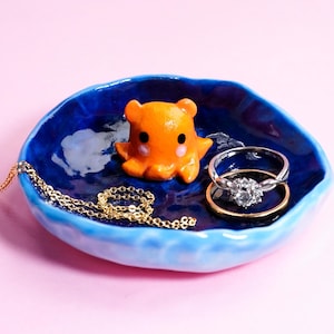 Può includere: Un vassoio blu in ceramica per anelli con una statuina di polpo arancione, una collana d'oro e due anelli. Il vassoio ha una finitura lucida e un bordo ondulato. Un anello ha una pietra grande e trasparente.