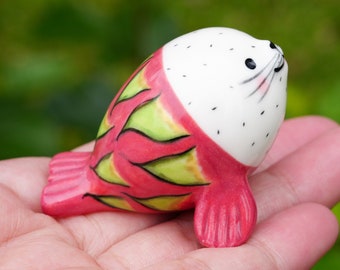 May include: A ceramic seal figurine with a white body and a pink and green dragon fruit design. The seal is sitting on its belly with its flippers outstretched.