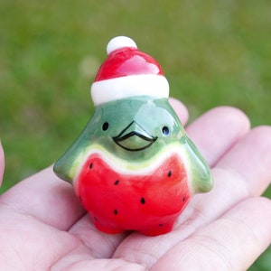 May include: A small ceramic figurine of a watermelon wearing a red and white Santa hat. The figurine is painted with green, red, and black details.