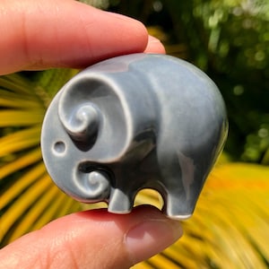 May include: A small, grey ceramic elephant figurine with a swirl design on its head. The figurine is approximately 5 cm tall and is being held in a person's hand.