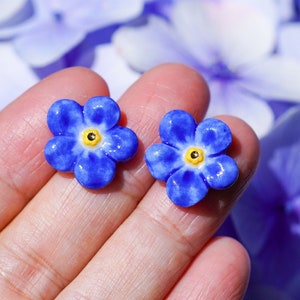 May include: A pair of blue forget-me-not flower earrings with yellow centers. The earrings are made of ceramic and are shown on a hand.