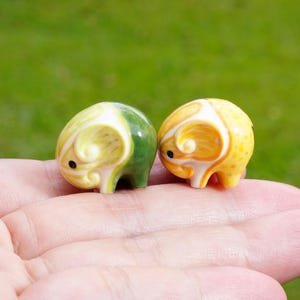 May include: Two miniature ceramic ram figurines. One is yellow and green, the other is yellow and orange. Both have a curled horn design and small black eyes. The figurines are held in a person's hand against a blurred green background.