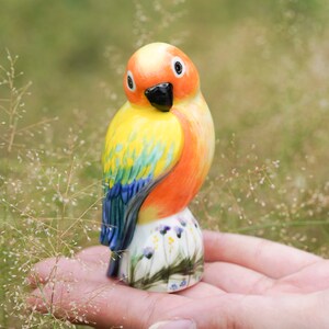 Könnte beinhalten: Eine Keramikfigur eines Papageis mit orangefarbener, gelber, blauer und grüner Farbe. Der Papagei sitzt auf einem Sockel mit einem bemalten Blumendesign.