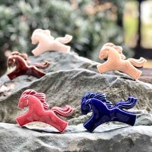 May include: Five ceramic horse figurines in various colors, including pink, brown, red, and blue. The figurines are arranged on a rock surface.