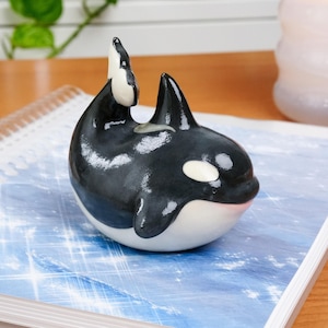 May include: A ceramic orca figurine, predominantly black and white, with a rounded body and upright dorsal fin. The orca has a white patch around its eye and a pink mouth. The figurine is displayed on a blue surface with a subtle pattern.