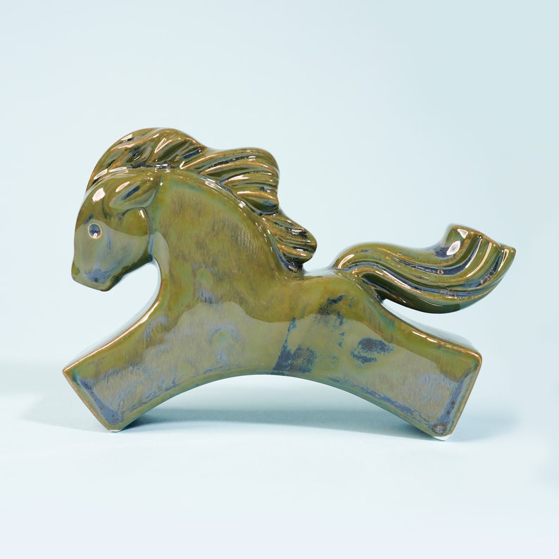 May include: A green ceramic horse figurine with a flowing mane and tail. The horse is in a running pose with its head tilted down.