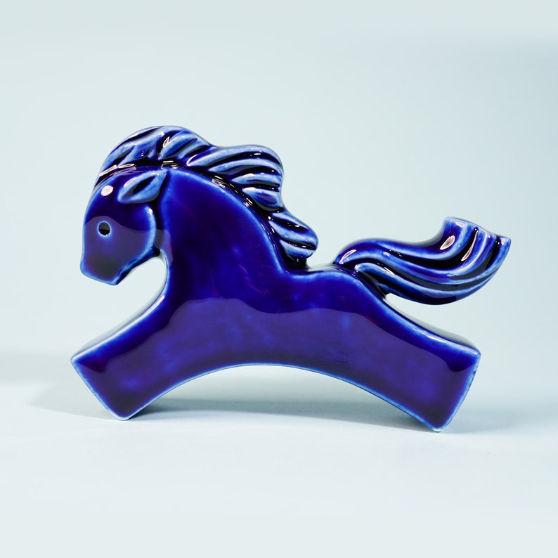 May include: A cobalt blue ceramic horse figurine with a stylized mane and tail. The horse is in a running pose with its head turned to the side.