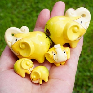 Puede incluir: Colección de figuritas de elefantes de cerámica amarilla con detalles en color marfil. Los elefantes varían en tamaño, el más grande mide aproximadamente 7.6 cm de largo. Las figuritas tienen un acabado brillante y están dispuestas en una mano.