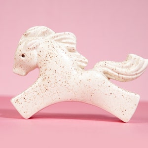 May include: A white ceramic horse figurine with brown speckles, running with its head turned to the side. The figurine has a long tail and mane.