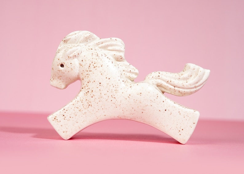 May include: A white ceramic horse figurine with brown speckles, running with its head turned to the side. The figurine has a long tail and mane.