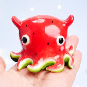 May include: A ceramic octopus figurine with a watermelon design. The red body has black seed details and white spots. The tentacles have green and white stripes. The eyes are large and black. The figurine is held in a hand.