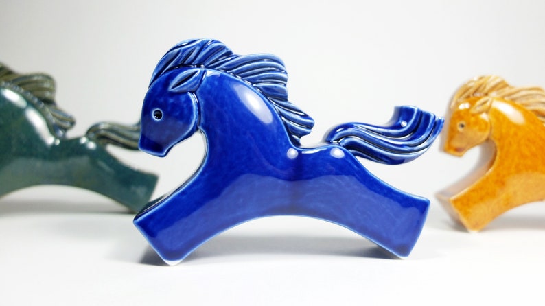 May include: Three ceramic horse figurines in different colors: blue, green, and orange. The blue horse is in the foreground and is facing the right. The green horse is behind the blue horse and is facing the left. The orange horse is behind the green horse and is facing the right.