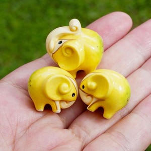 May include: Three small, yellow ceramic ram figurines with cream-coloured horns and facial details. The rams have a rounded body shape and are posed on a hand, with a green background visible.