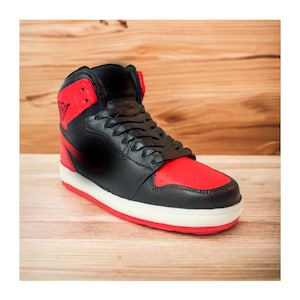 Puede incluir: Una zapatilla alta negra y roja con suela blanca. La zapatilla presenta un logotipo negro y rojo en el lateral. Los cordones son negros y el fondo es de madera.