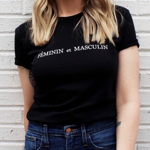 Puede incluir: Una camiseta negra con el texto "FEMININ et MASCULIN" en letras blancas. La persona lleva pantalones vaqueros azules.