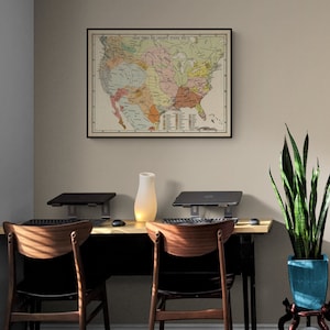 May include: A vintage map of the United States showing the language groups of Native American tribes, framed in black and hanging on a beige wall. The map is surrounded by a desk with two chairs, a potted snake plant, and a lamp.