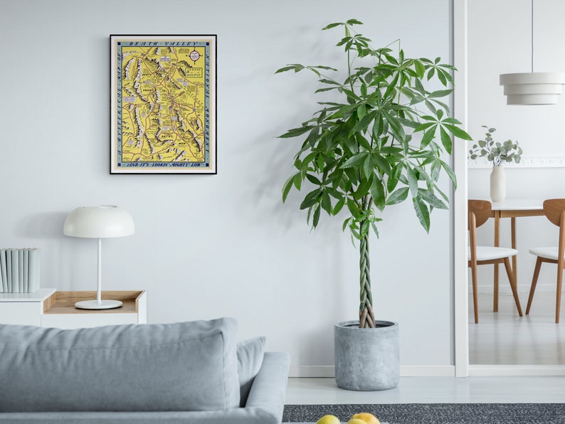 May include: A framed vintage map of Death Valley, California, with a yellow background and blue and black text. The map is titled "Death Valley and Its Surrounding Country" and is dated "1907". The map is hanging on a white wall in a modern living room.