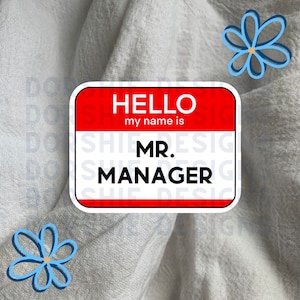May include: A red and white name tag with the text "HELLO my name is MR. MANAGER" on a white fabric background with blue flowers.