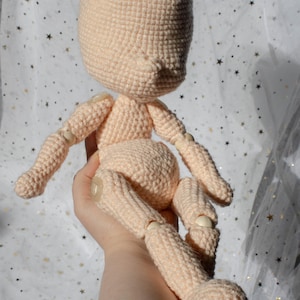 Puede incluir: Un cuerpo de muñeca de ganchillo color melocotón con extremidades articuladas. La cabeza, los brazos y las piernas de la muñeca están conectados con pequeñas articulaciones de color claro. La muñeca se sostiene en una mano sobre un fondo de tela blanca con estrellas doradas.