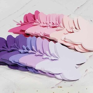 Puede incluir: Un mont&oacute;n de mariposas de papel superpuestas en tonos de morado, rosa y blanco. Las mariposas est&aacute;n dispuestas en forma de abanico, con la mariposa m&aacute;s grande en la parte inferior y la m&aacute;s peque&ntilde;a en la parte superior.