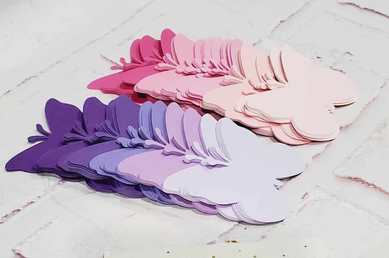 Puede incluir: Un mont&oacute;n de mariposas de papel superpuestas en tonos de morado, rosa y blanco. Las mariposas est&aacute;n dispuestas en forma de abanico, con la mariposa m&aacute;s grande en la parte inferior y la m&aacute;s peque&ntilde;a en la parte superior.