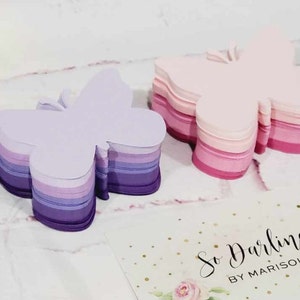 Puede incluir: Dos pilas de notas de papel con forma de mariposa. La pila de la izquierda es morada, lavanda y blanca. La pila de la derecha es rosa y blanca. Las pilas est&aacute;n sobre una superficie blanca con una tarjeta de visita que dice "So Darling by Marisol".