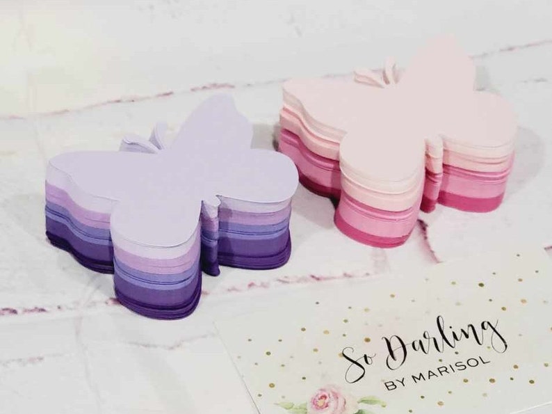 Puede incluir: Dos pilas de notas de papel con forma de mariposa. La pila de la izquierda es morada, lavanda y blanca. La pila de la derecha es rosa y blanca. Las pilas est&aacute;n sobre una superficie blanca con una tarjeta de visita que dice "So Darling by Marisol".