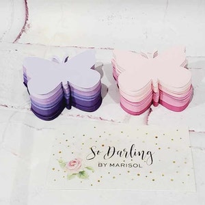 Puede incluir: Dos pilas de recortes de mariposas de papel superpuestas. La pila de la izquierda es morada y lavanda, la pila de la derecha es rosa y roja. Una tarjeta blanca con purpurina dorada y el texto "So Darling by Marisol" est&aacute; en primer plano.