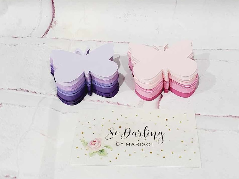 Puede incluir: Dos pilas de recortes de mariposas de papel superpuestas. La pila de la izquierda es morada y lavanda, la pila de la derecha es rosa y roja. Una tarjeta blanca con purpurina dorada y el texto "So Darling by Marisol" est&aacute; en primer plano.