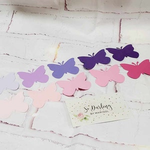 Puede incluir: Surtido de mariposas de papel en varios tonos de morado, rosa y lavanda. Las mariposas est&aacute;n recortadas y dispuestas sobre un fondo de ladrillo blanco. Una peque&ntilde;a tarjeta con el texto "So Darling by Marisol" se encuentra en la esquina inferior derecha.