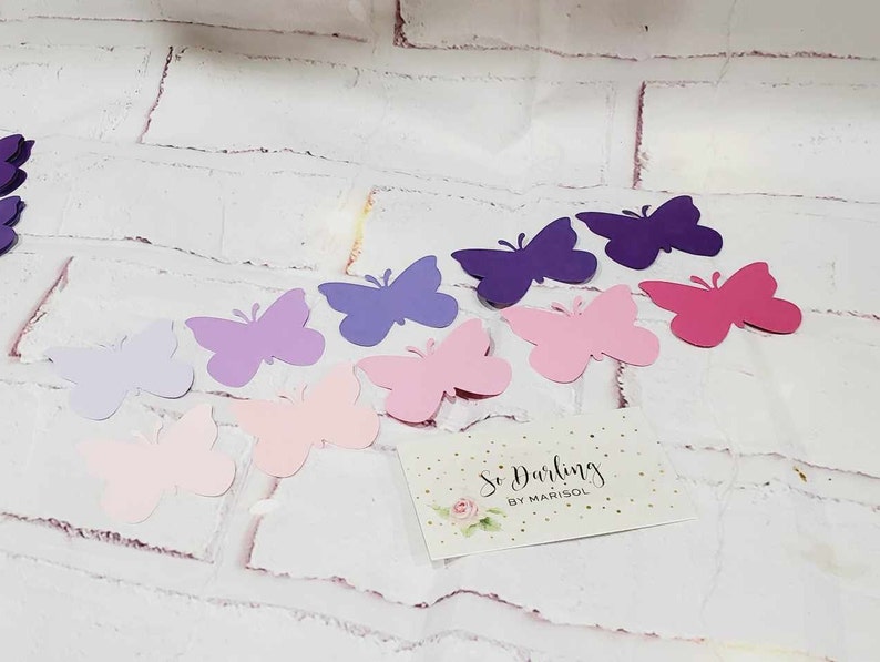 Puede incluir: Surtido de mariposas de papel en varios tonos de morado, rosa y lavanda. Las mariposas est&aacute;n recortadas y dispuestas sobre un fondo de ladrillo blanco. Una peque&ntilde;a tarjeta con el texto "So Darling by Marisol" se encuentra en la esquina inferior derecha.