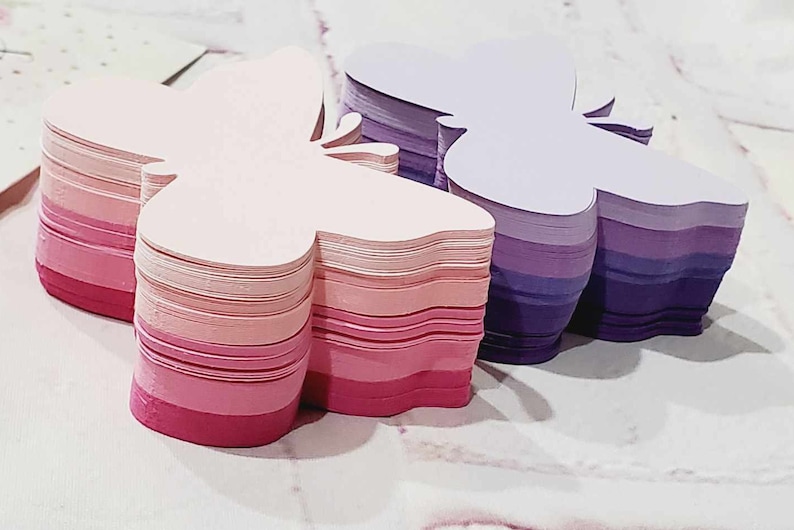 Puede incluir: Dos pilas de notas de papel con forma de mariposa. La pila de la izquierda es rosa y blanca, mientras que la pila de la derecha es morada y blanca.
