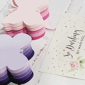 Puede incluir: Dos pilas de recortes de papel con forma de mariposa. La pila superior es rosa y la pila inferior es morada. Las pilas est&aacute;n sobre una superficie blanca. Una tarjeta blanca con purpurina dorada y el texto "So Darling by Marisol" est&aacute; en el fondo.